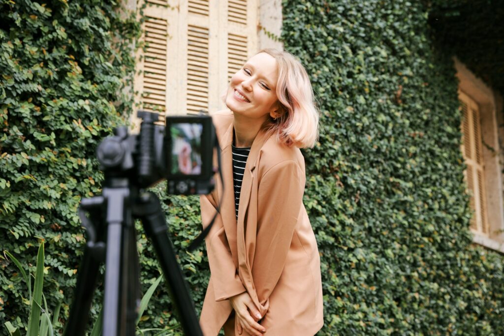 Light skinned female standing in front of a greenery covered collage in front of a camera recording herself. She is wearing a peach blazer and black and white striped shirt. 